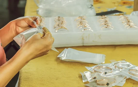 Close-up of a worker carefully packaging gold-plated clover bracelets into individual protective poly bags at a jewelry manufacturing factory.